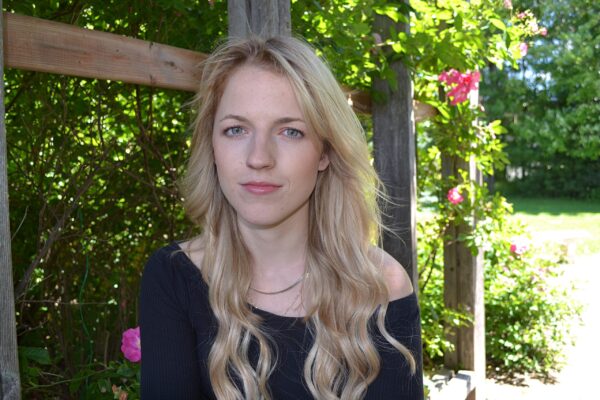 一位金发长发、身穿深蓝色上衣的年轻女子坐在一个长满绿色植物和粉色花朵的花园前，一脸严肃地盯着镜头。