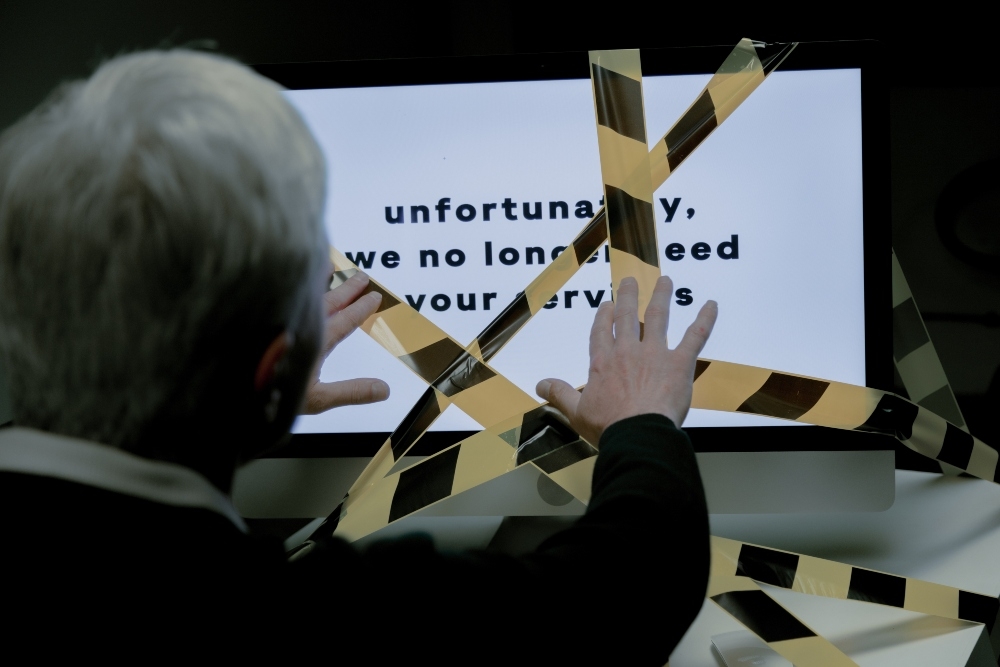 一名头发花白、戴着眼镜的男子把手举到电脑屏幕前，电脑屏幕上贴着黄黑相间的警用胶带。屏幕上的文字写着，很不幸，我们不再需要您的服务。