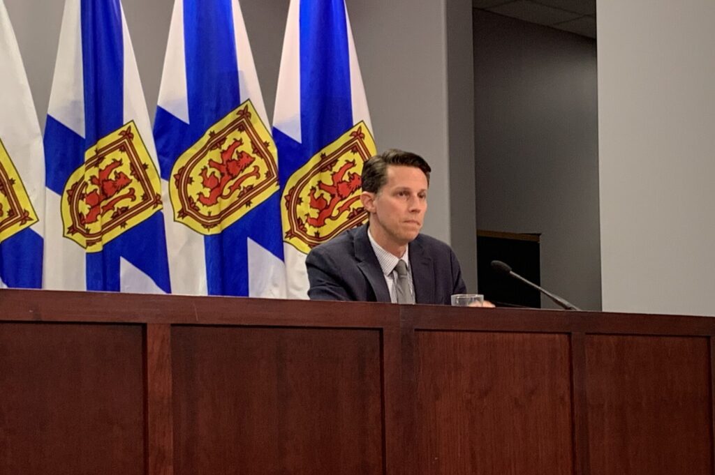 一个西装革履的男人坐在麦克风前。他身后是一排新斯科舍省的国旗——白、蓝、黄、红四色。