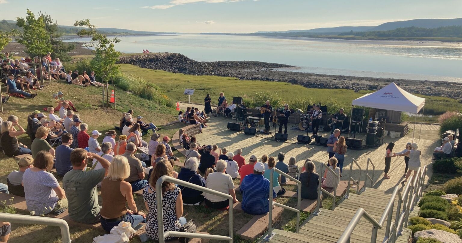 一小群人坐在户外的台阶上，俯瞰着一片水域。一支乐队在人群前表演，一对夫妇在跳舞。