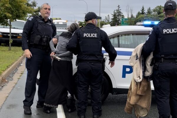 三名警察和一名戴着手铐的老妇人站在警车前。她拿着一根羽毛。