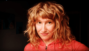 一个留着齐肩长红色波浪头的女人，穿着一件橙色拉链卫衣。