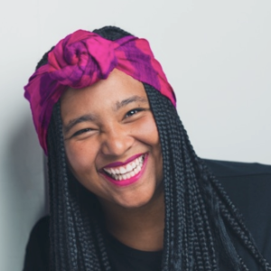 一个年轻的黑人女性，梳着长长的辫子，戴着亮粉色的头巾和黑色上衣，笑容灿烂