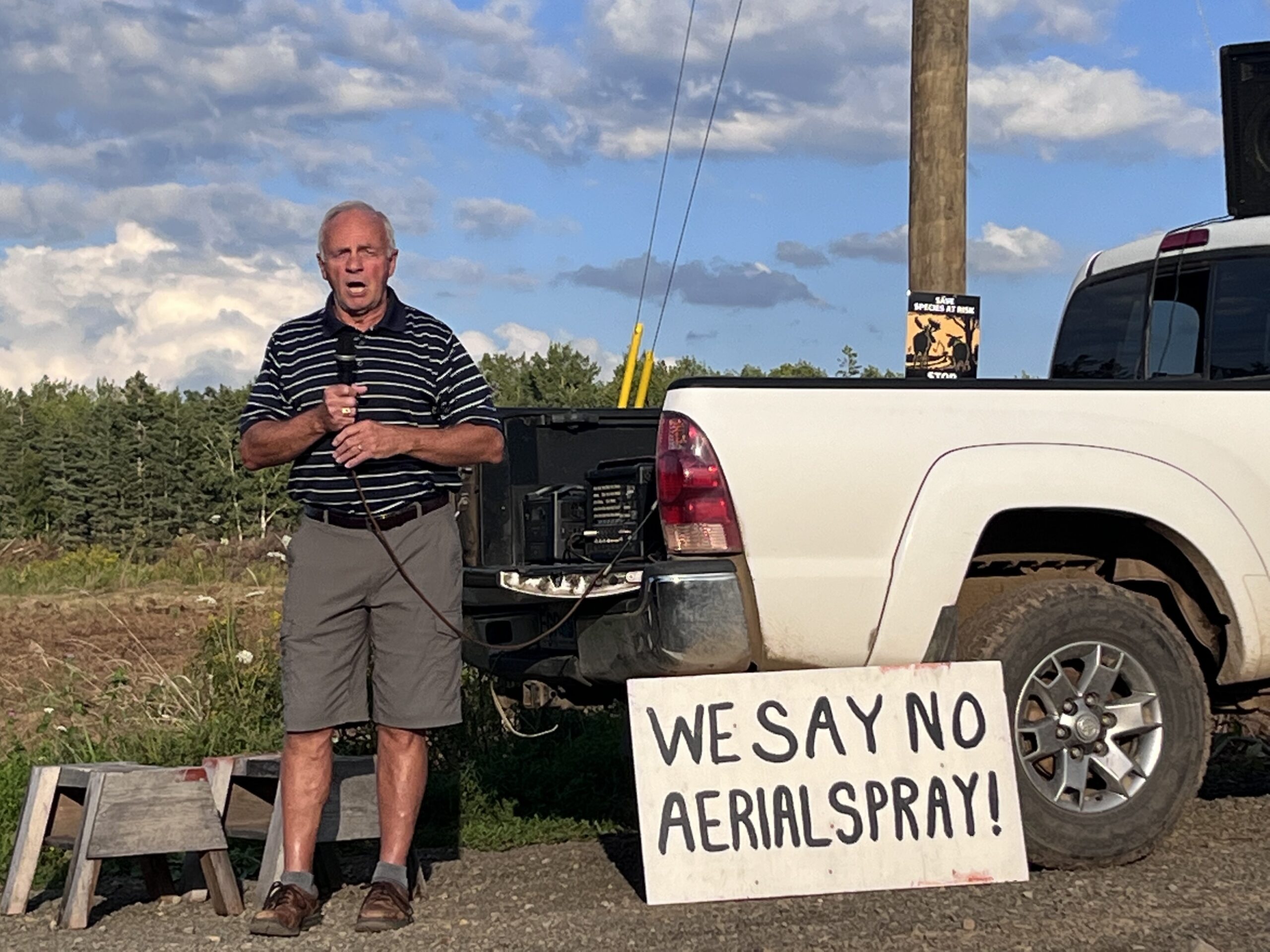 一名中年白人男子对着田野边一辆皮卡旁的麦克风说话。他旁边的牌子上写着“我们对空中喷雾剂说不”。