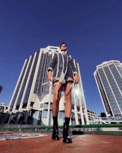 一名男子穿着蓝色镶边和流苏的夹克，脚上穿着黑色靴子，在两座海滨办公大楼前摆姿势。