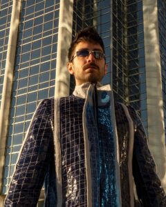 一名男子身穿蓝色镶边和流苏外套，脚穿黑色靴子，在两座海滨办公大楼前摆姿势。