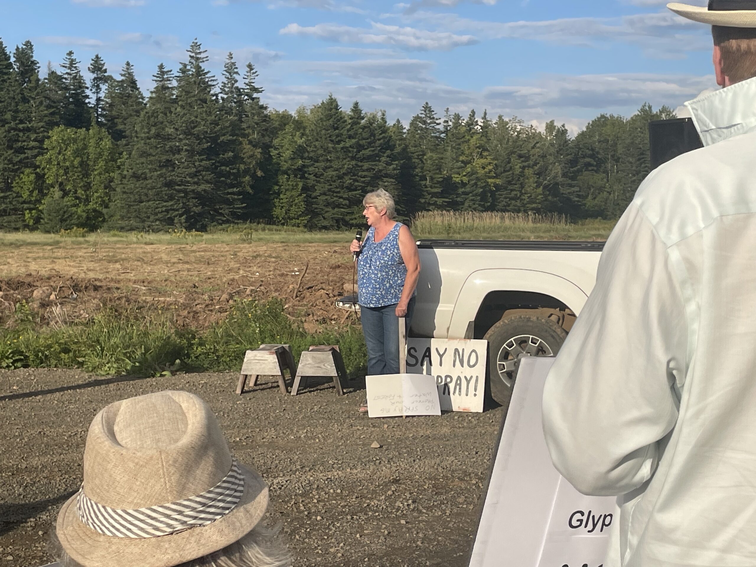 一名中年白人妇女在空旷的田野和一辆白色皮卡旁对着麦克风说话。前景中可以看到两名抗议者的背影