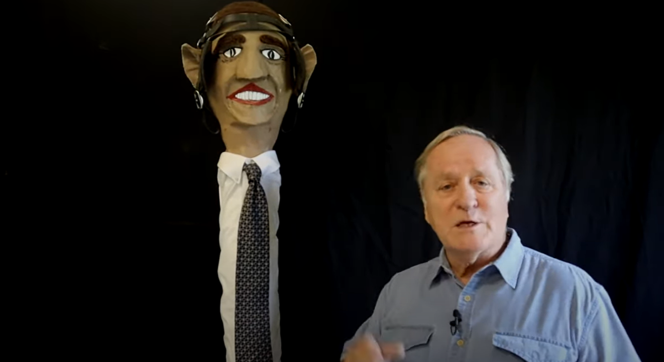 一名身穿蓝色衬衫的中年白人男子站在一尊西装男子木雕的旁边。