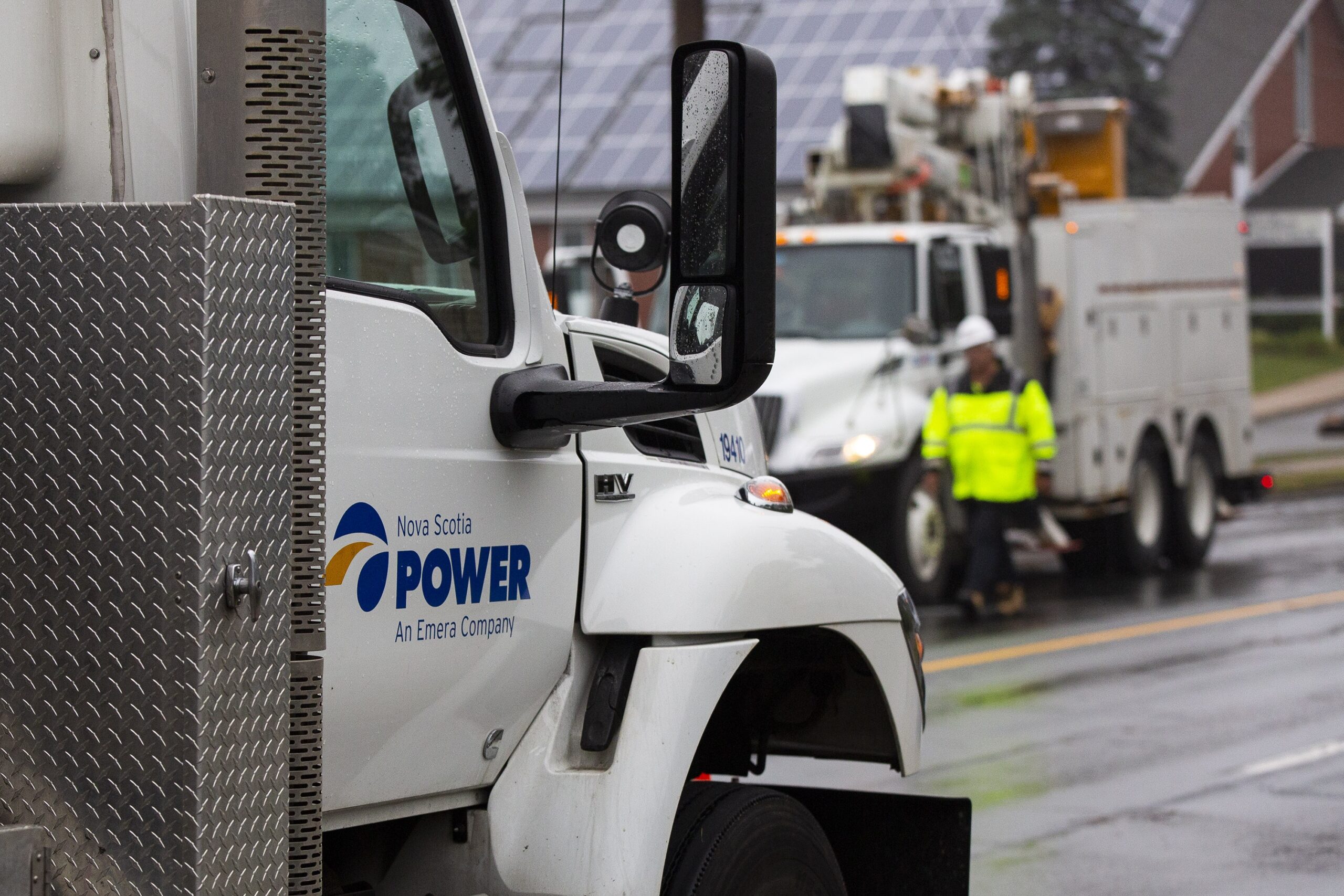 一辆带有蓝色新斯科舍省电力公司标志的卡车出现在前景中。在它后面，一名身穿荧光黄色衣服的男子走过另一辆卡车。