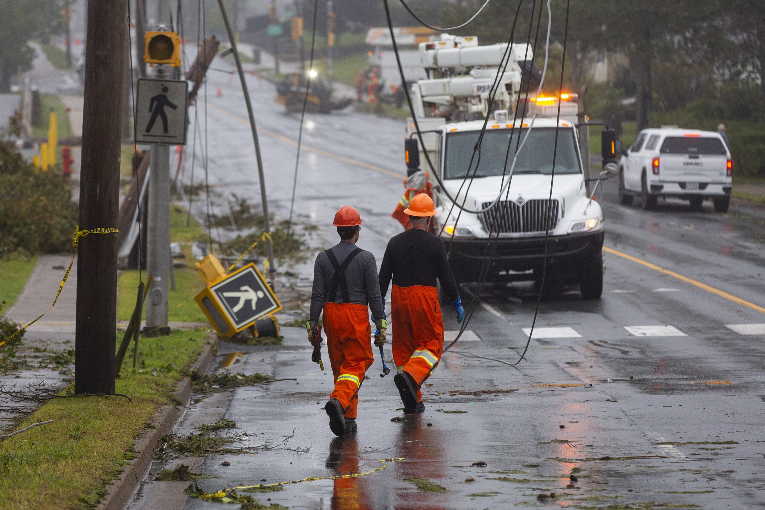 两名工人穿着橙色的裤子，戴着安全帽，走在一条凌乱的街道上，电线从上面垂下来，地上有一个步行标志。