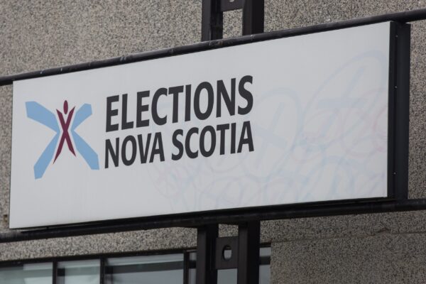 在一座灰色的建筑上，可以看到一个黑色金属管道框架的标志。它有一个浅蓝色盾徽中心的酒红色X标志，以及选举NOVA SCOTIA字样。