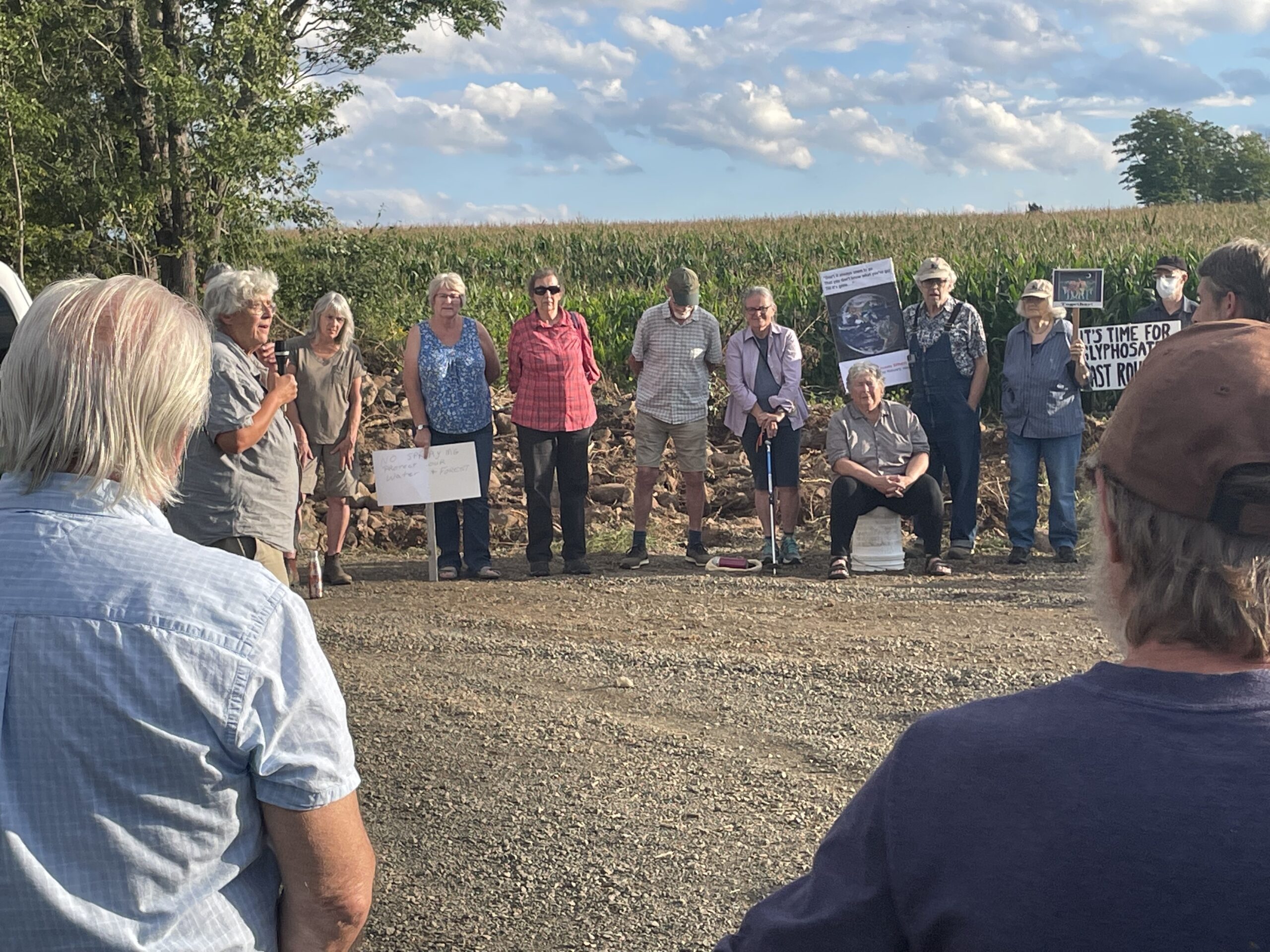 一个阳光明媚的日子，一位白发苍苍的妇女对聚集在玉米地外的人群讲话