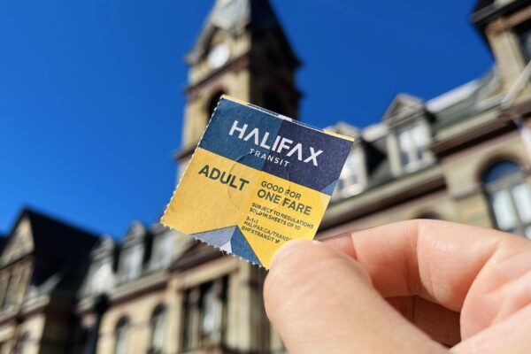一只手拿着一张蓝黄相间的哈利法克斯过境车票。在背景中，模糊的是一座古老的砂岩建筑。它是晴朗的。
