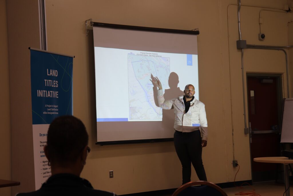 一个黑人站在屏幕前，向一群观众展示地图。屏幕旁边是一条蓝白相间的横幅，上面写着“土地所有权倡议”