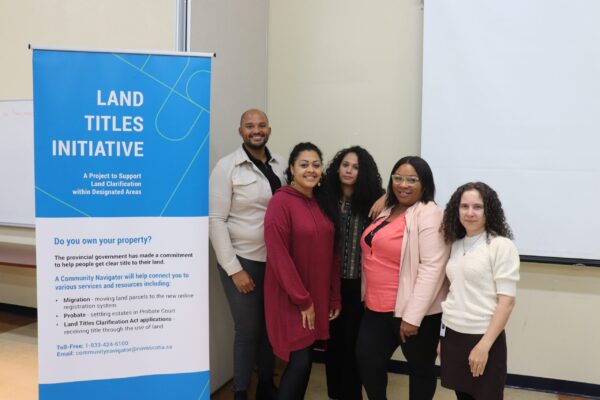 一名黑人男子和三名有色人种妇女站在一块蓝白相间的牌子旁，牌子上写着“土地所有权倡议”，这是一个支持在指定区域内澄清土地的项目。