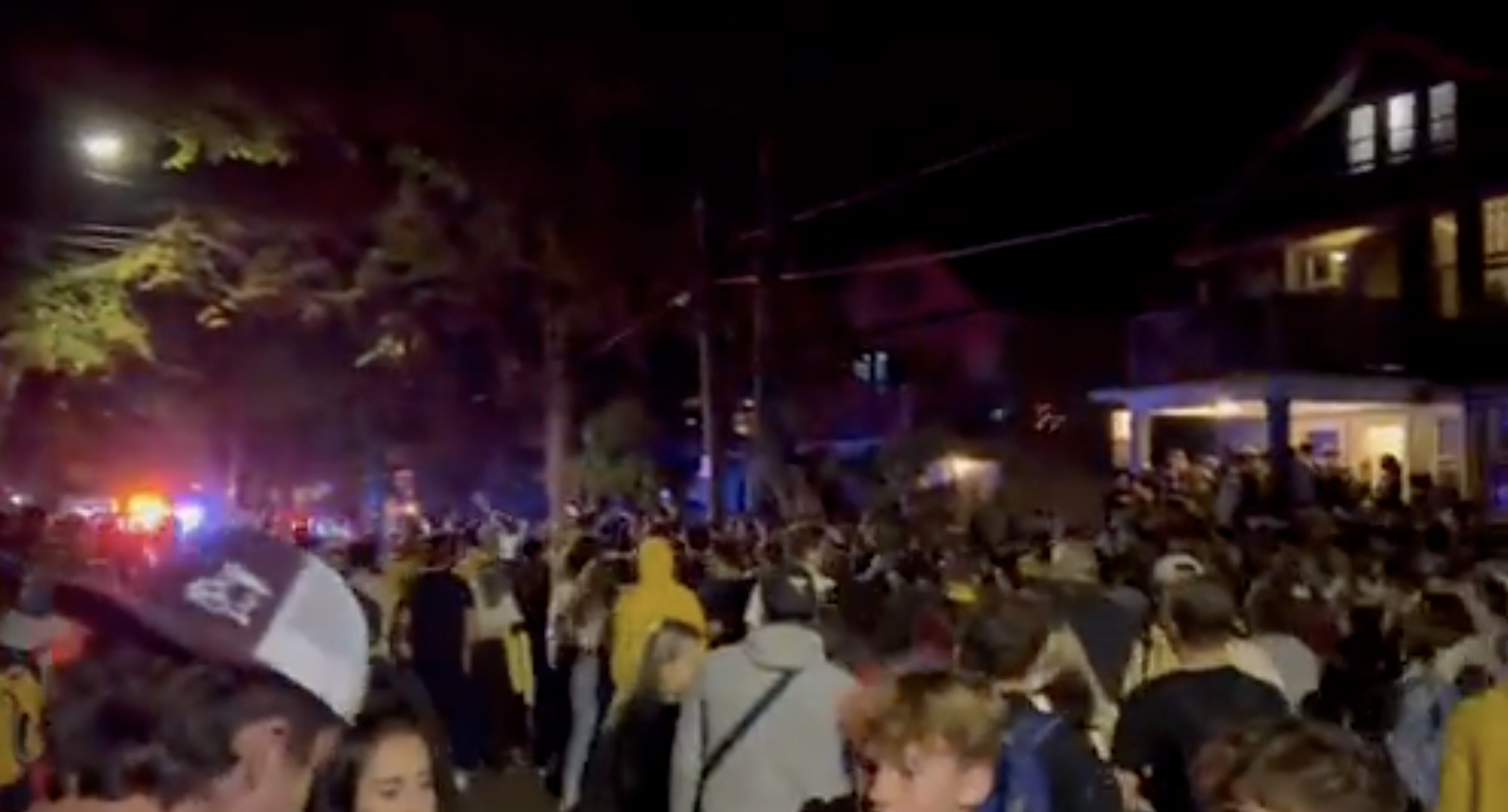 晚上，街道上挤满了参加聚会的学生。警灯在背景中刺眼。一幢住宅在人群的一侧。