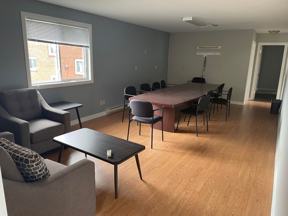 一个有会议室桌子和椅子的大房间。角落里有一把椅子，一个小沙发和一张咖啡桌。远处墙上的窗户让光线照进来。