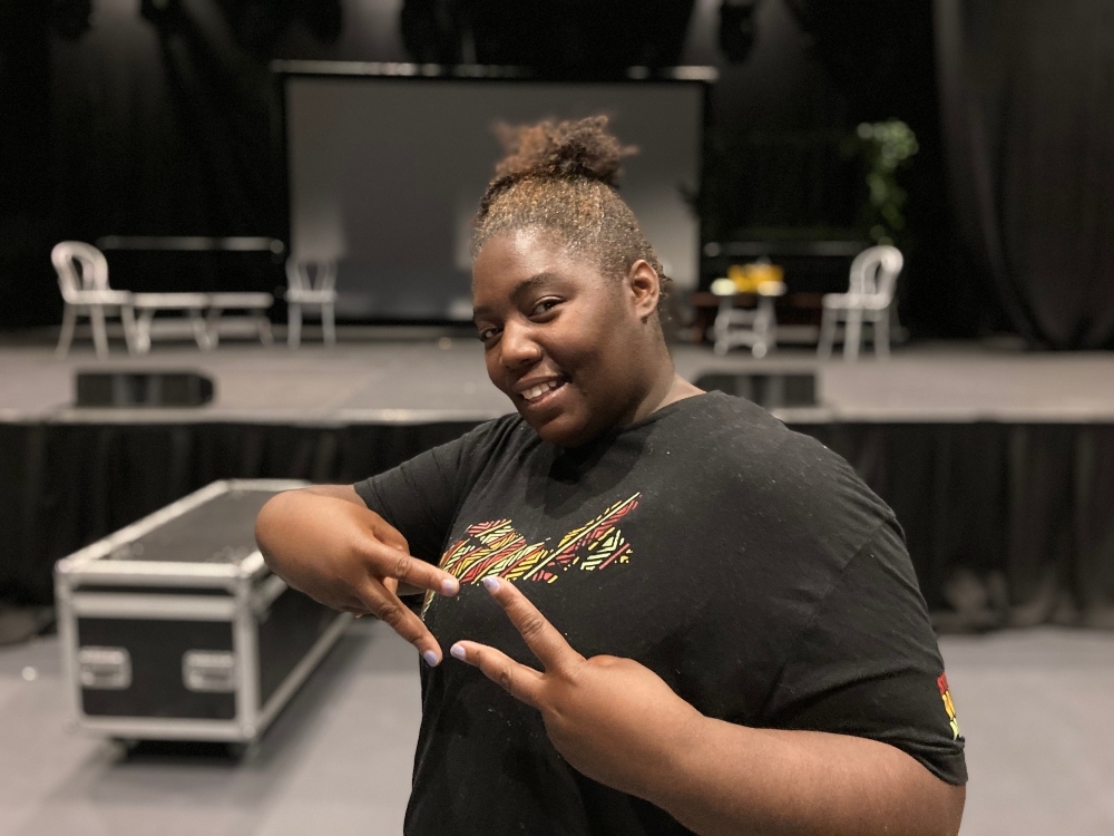 一位身穿黑色t恤、扎着头发的黑人妇女站在剧院舞台前，打着两个和平手势