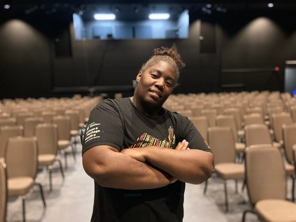 一名身穿黑色t恤、头发扎起来的黑人妇女抱着双臂站在一间放着一排排米色椅子的空荡荡的剧院里。