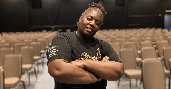 一名身穿黑色t恤、头发扎起来的黑人妇女抱着双臂站在一间放着一排排米色椅子的空荡荡的剧院里。