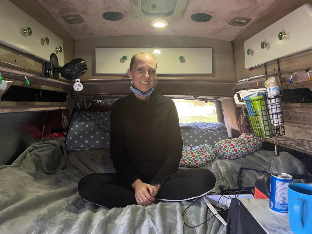 一名浅棕色头发、下巴下戴着口罩的白人妇女坐在一辆货车内的床上，脸上涂着苹果酱。