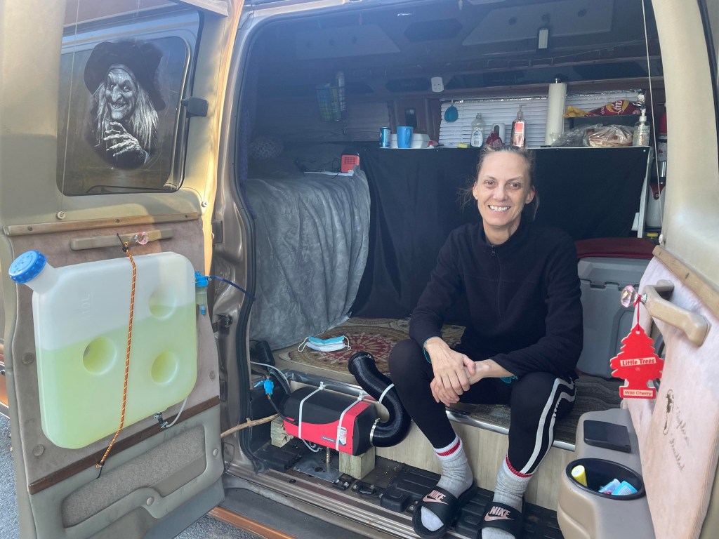 一名浅棕色头发向后梳，穿着黑色毛衣和打底裤的白人妇女就坐在一辆棕金色面包车里。里面有一张床，一个带有水槽和食物的台面。门上有一个柴油加热器和一个女巫的万圣节大贴纸。