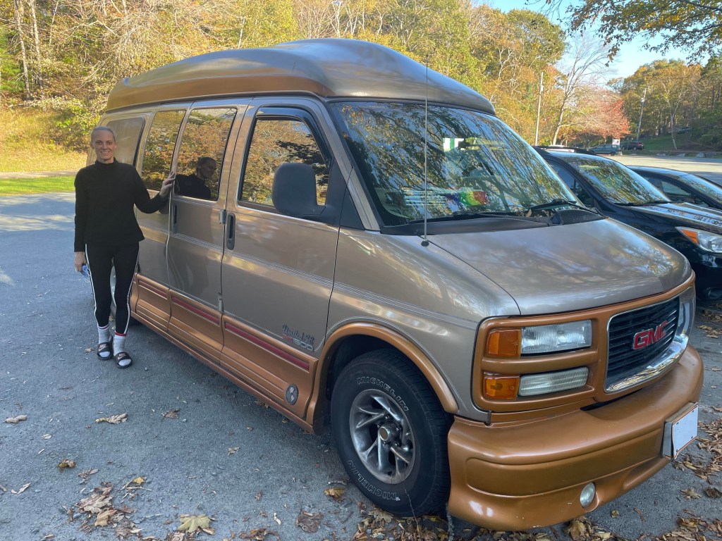 一名身穿黑色外套、袜子和人字拖的白人女子站在停车场的一辆金棕色面包车外。地上有几片树叶