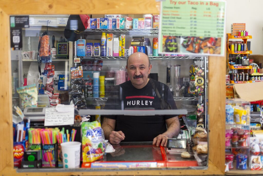 一名留着小胡子、身穿黑色t恤的男子站在有机玻璃盾牌后面的柜台前，周围摆满了商品。