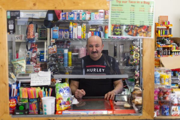 一名留着小胡子、身穿黑色t恤的男子站在有机玻璃盾牌后面的柜台前，周围摆满了商品。