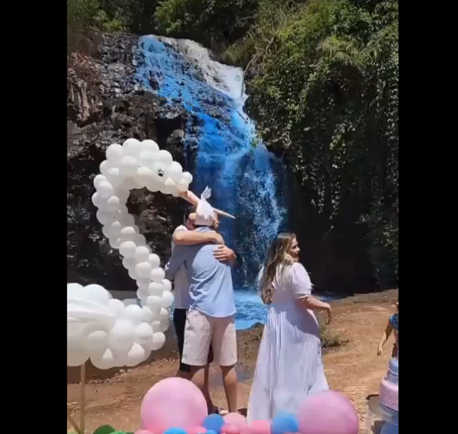 一个男人拥抱另一个男人，一个穿着白色长裙的女人站在旁边。有一只由白色气球和粉色和蓝色气球做成的鹳漂浮在地面上。背景是染成蓝色的瀑布。