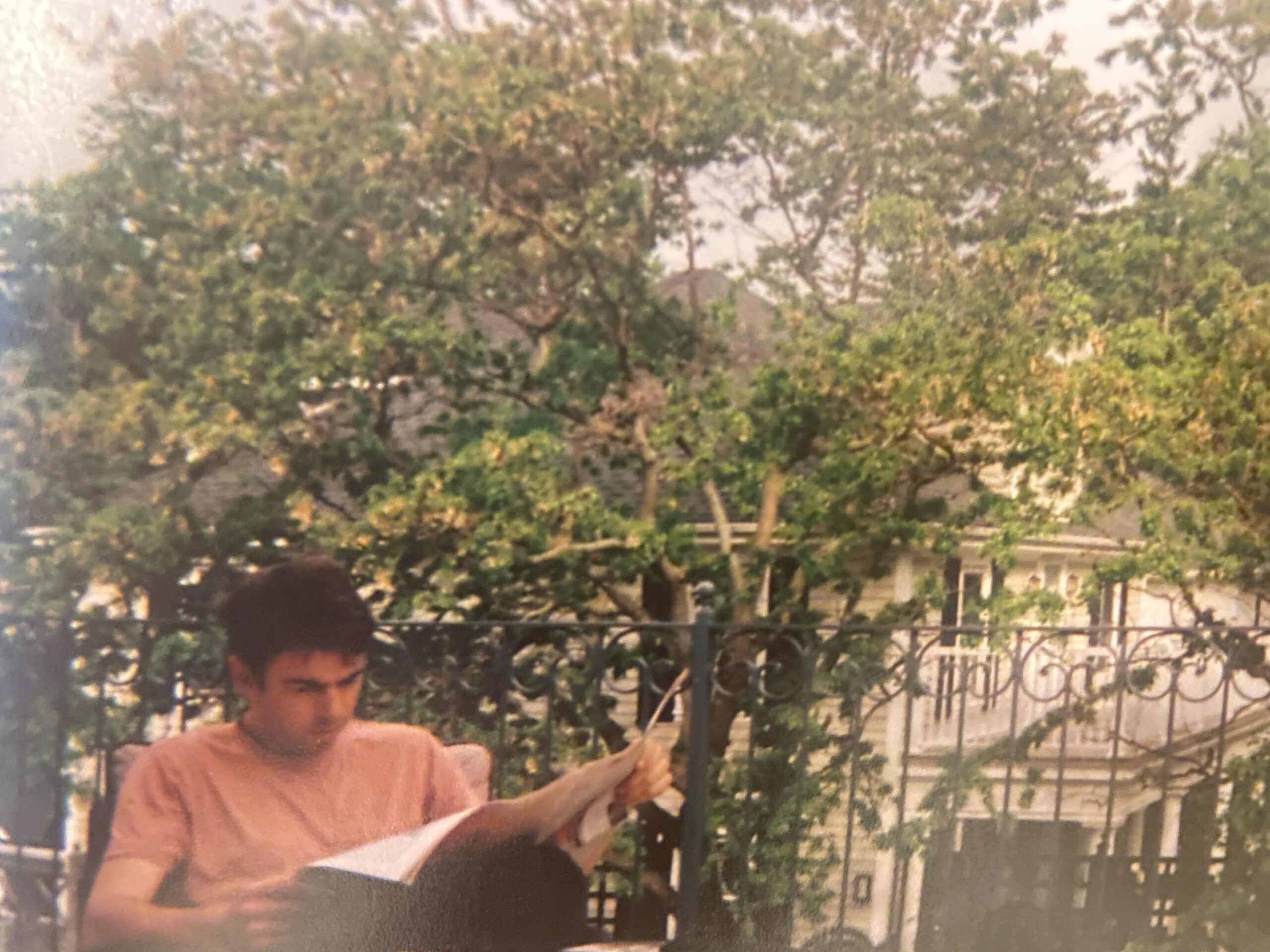 一个穿粉红色衬衫的年轻人坐在那里，膝上放着一张打开的报纸。这是一天的时间。他是在外面。树叶占据了整个背景。