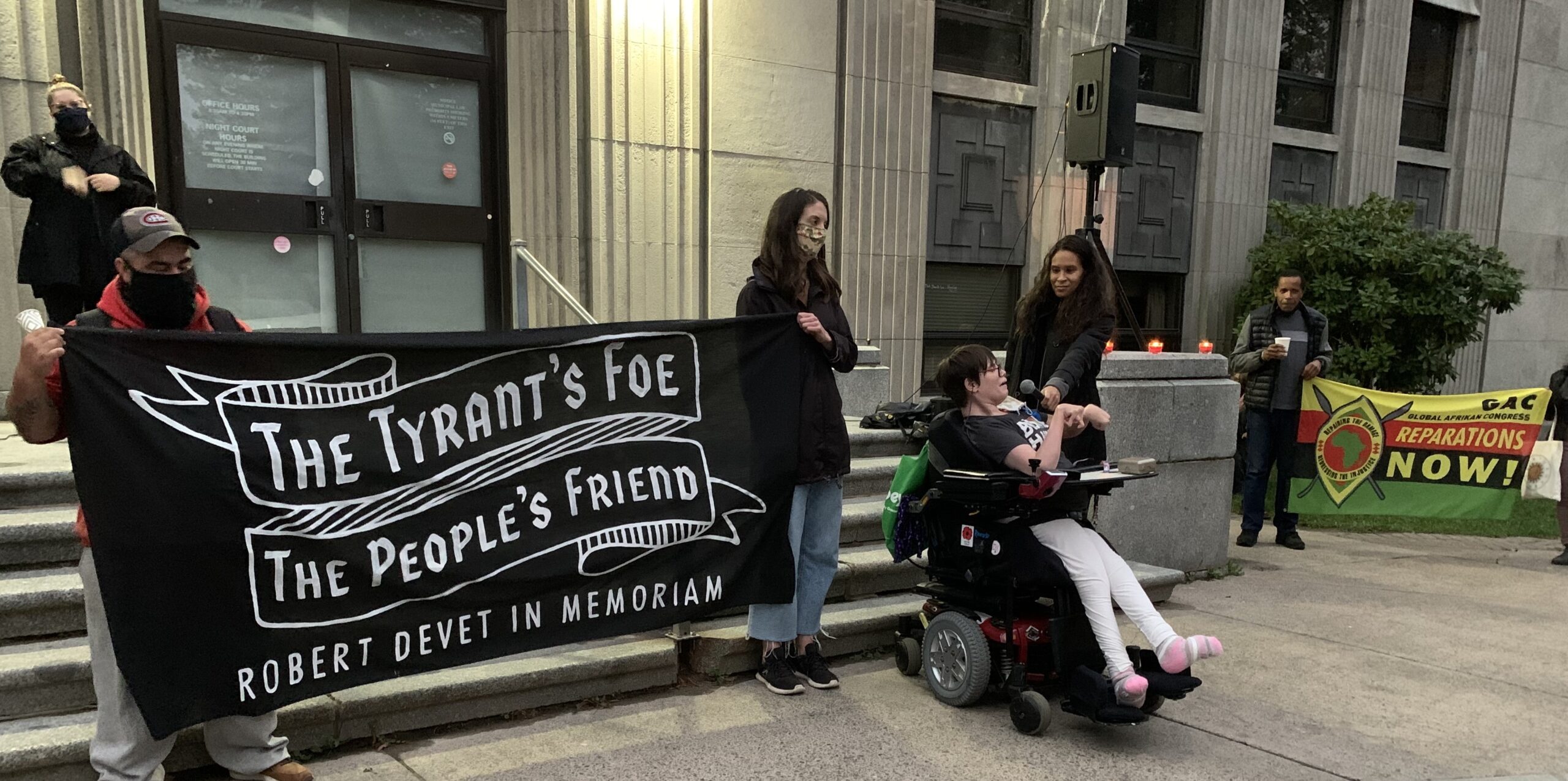 Robert Devet纪念馆的一张照片显示，两名戴着面具的人举着一块黑白相间的牌子，上面写着“暴君的敌人，人民的朋友，Robert Devet纪念馆”。艾尔·琼斯正拿着麦克风，以便坐轮椅的活动人士维姬·里瓦克(Vicky Levak)讲述德维特和他的工作。