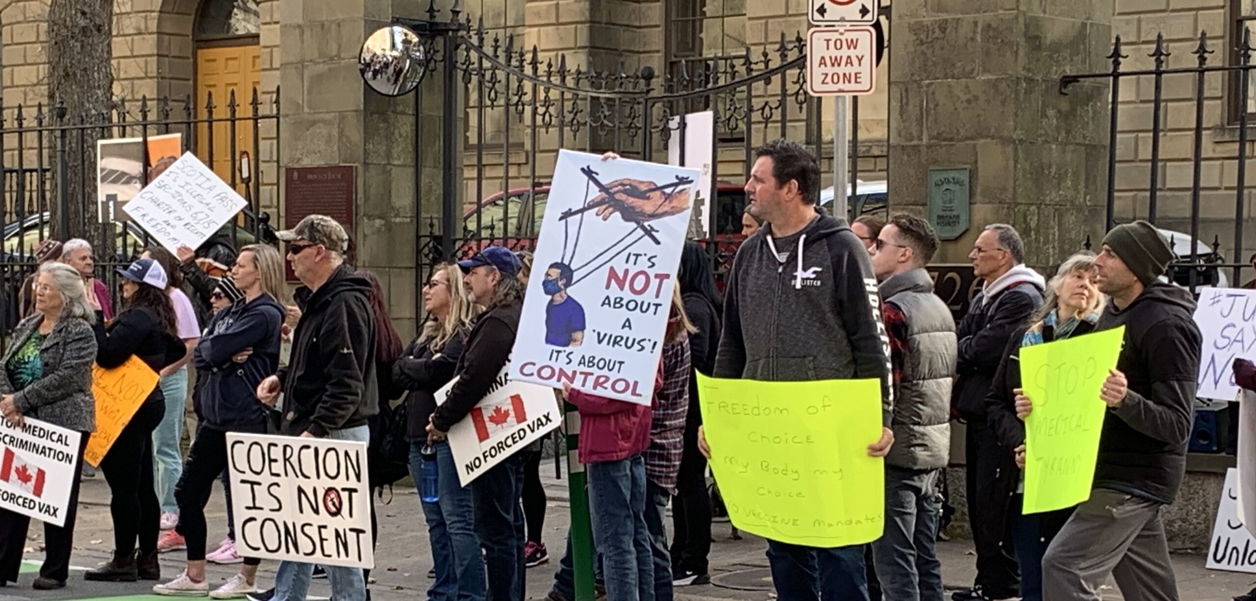 霍利斯街省政府大楼外的反疫苗抗议者的照片。抗议者举着手工制作的标语，上面写着强迫不是同意，这与病毒无关，而是控制。