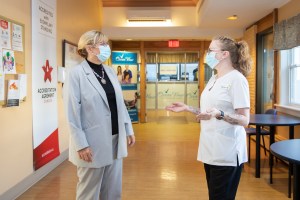 一张老年人和长期护理部长芭芭拉·亚当斯与注册护士香农·莫里斯·哈顿在Ocean View Continuing Care设施会面的照片。两个女人都站在医院的走廊上聊天。