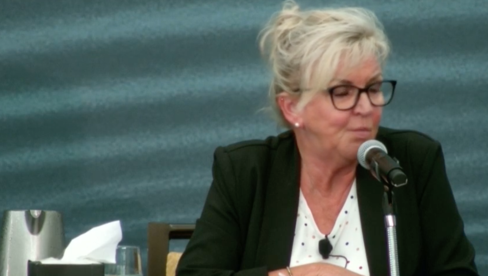 一个金发向后梳，白色上衣外穿黑色运动夹克的女人坐在麦克风前。